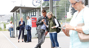 Zu sehen sind Menschen an einer Bushaltestelle die auf ihre Smartphones blicken. 